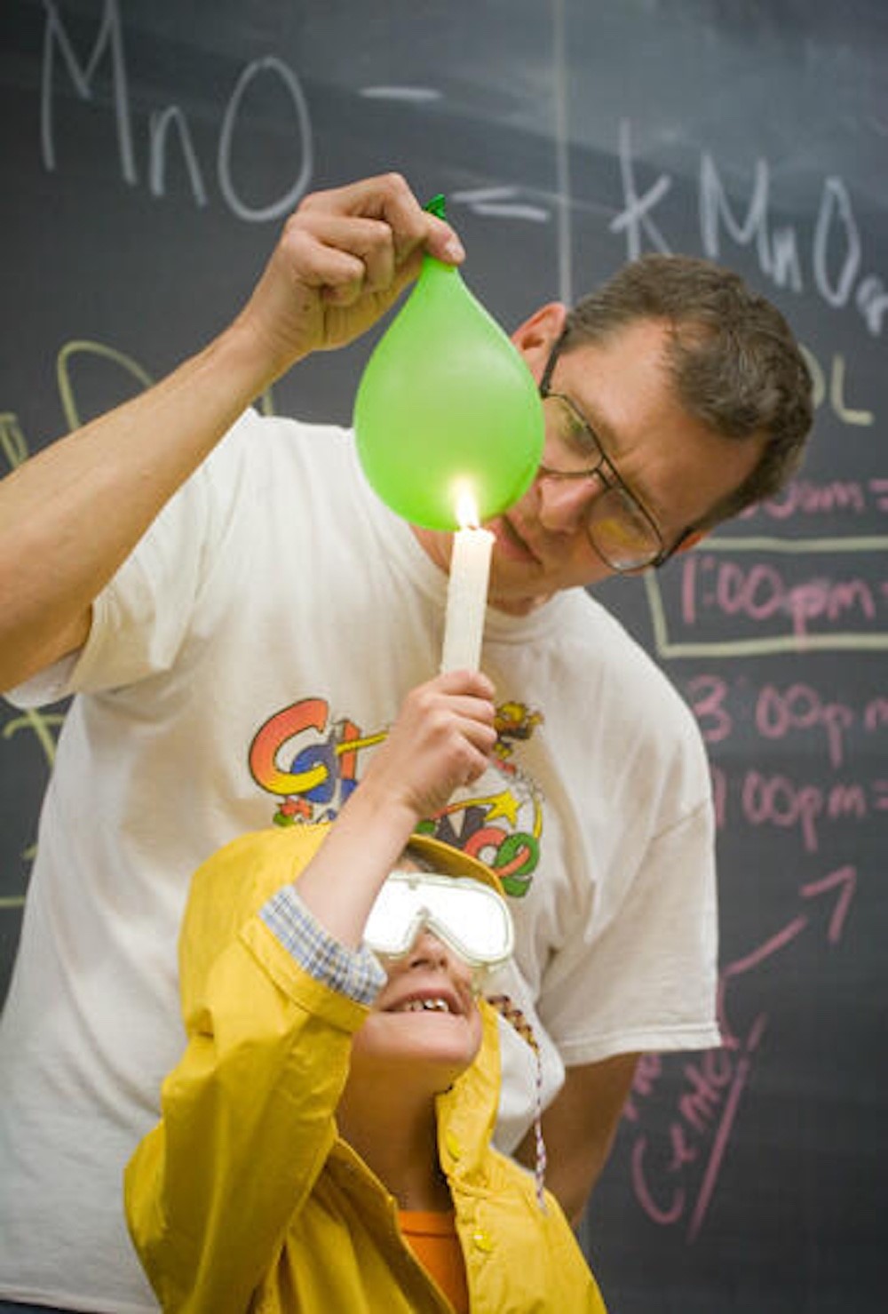 A man and a child stand in front of a blackboard. The man is holding a balloon, and the child is lifting a lit candle towards it.
