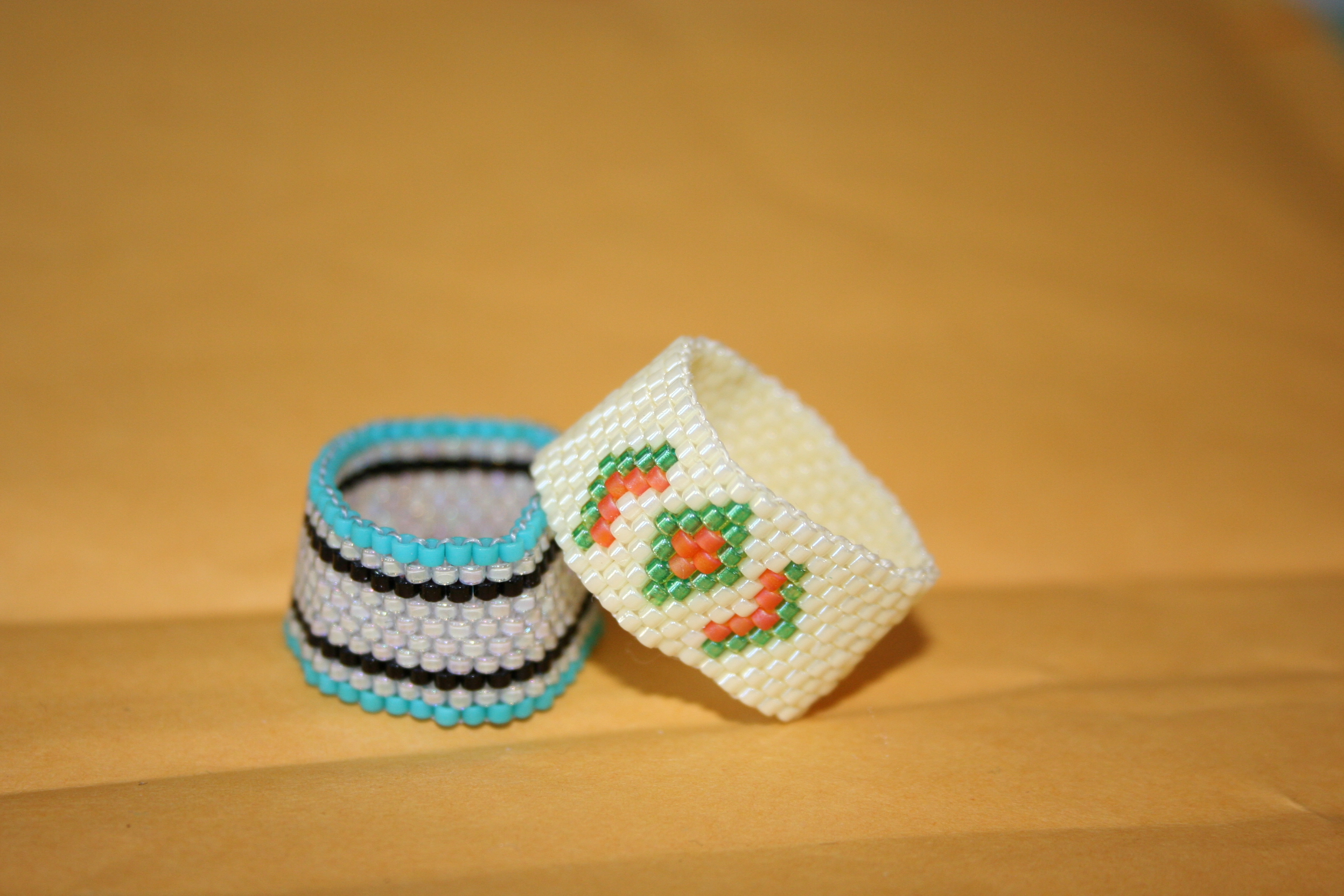 Image of two beaded rings made using peyote stitch. One is white with a green and orange geometric design, and the other has blue, black and white stripes.
