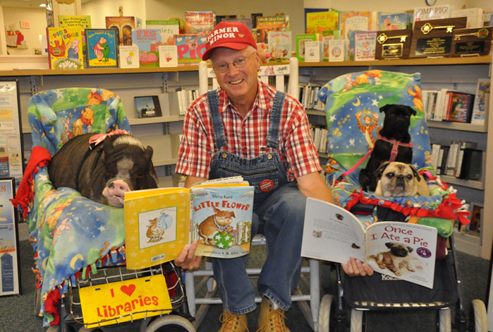 Farmer Minor sitting in chair and holding 2 books open for Daisy the pig and two pugs.