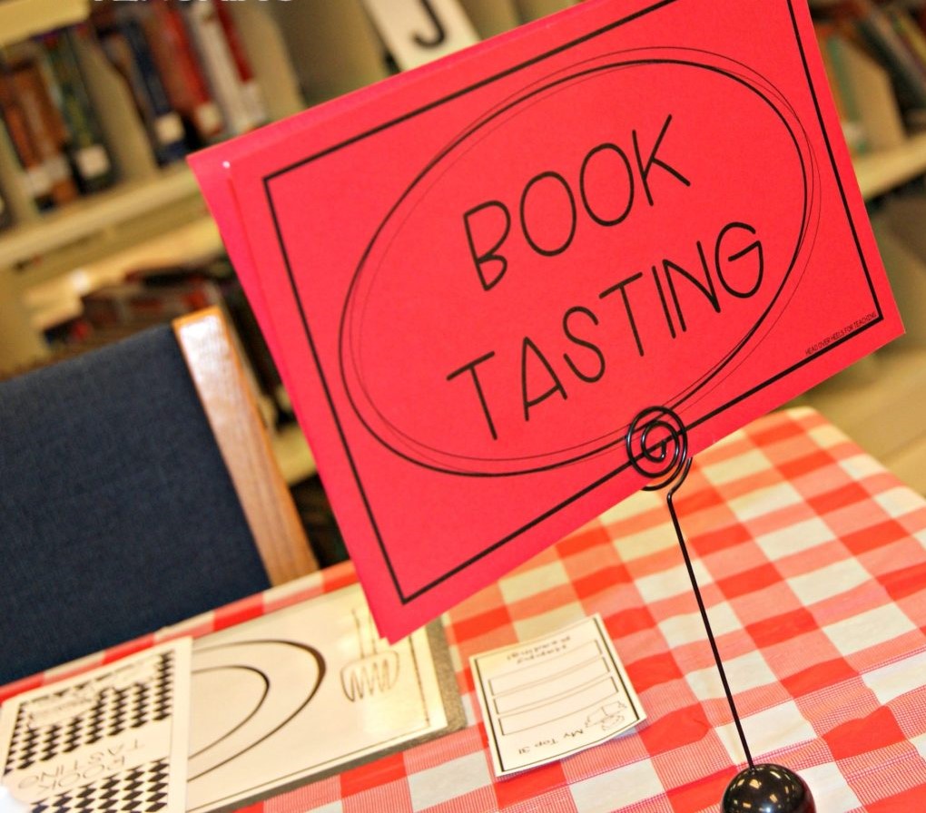 Table with a red gingham table cloth and a black place card holder with a red piece of paper that says "Book Tasting". There are library shelves full of books in the background.
