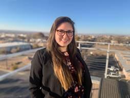 Noea Moss, a woman with long brown hair and glasses, stands outside smiling 